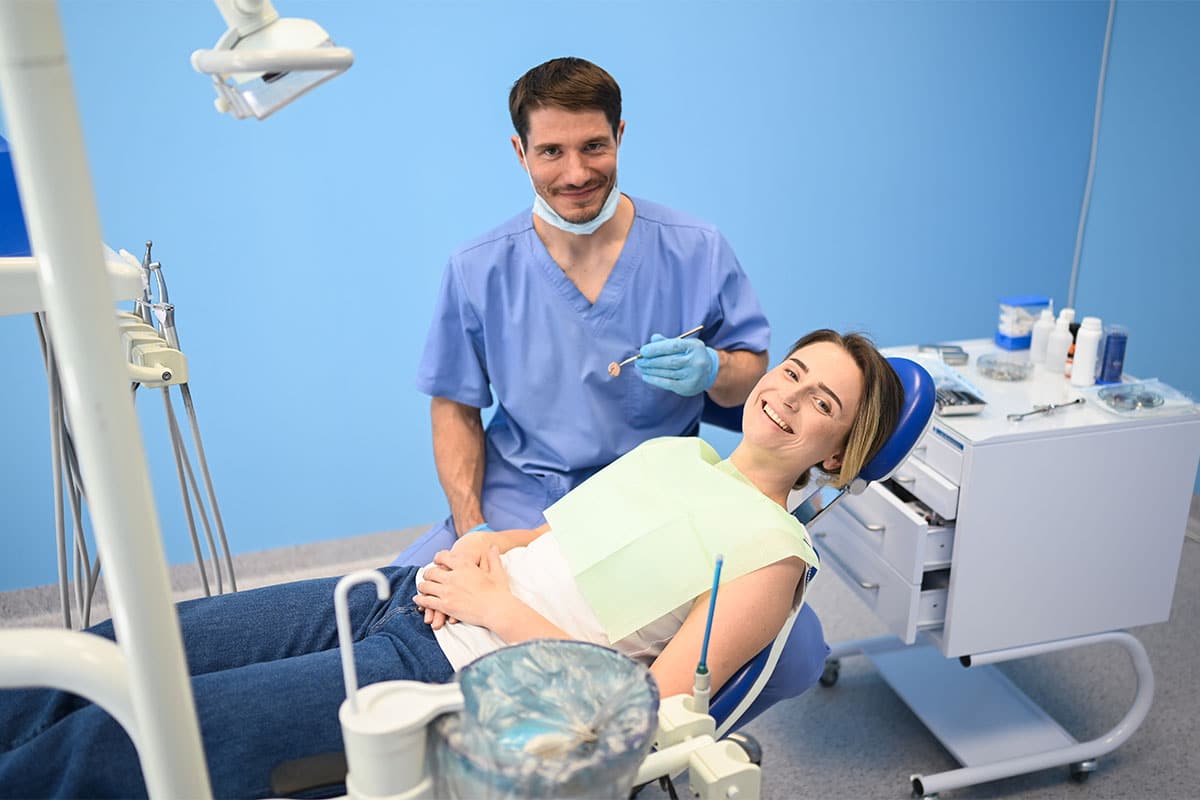 A dentist in blue scrubs smiles at the camera while holding a dental tool, standing next to a patient sitting in a dental chair, who is also smiling. The dental office setting is clean and brightly lit.