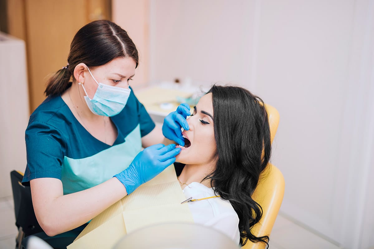 cosmetic dentistry | A dentist wearing a mask and gloves examines the teeth of a woman with dark hair, who is seated in a dental chair with a bib. The dentist uses dental tools inside the patients mouth.
