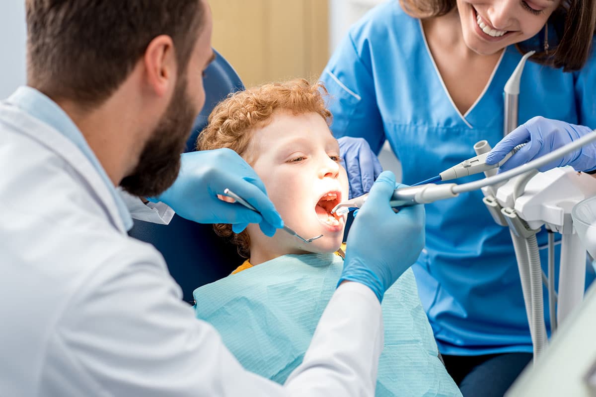 A young child sits in a dental hygiene care chair with their mouth open as a dentist examines their teeth. A dental assistant, both wearing blue gloves, assists and smiles beside them. Blog info page - Emergency Dentist Services in North Oakville